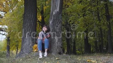 美妙的秋景。 迷人的年轻快乐微笑的女人坐在公园的草地上，在笔记本上做笔记。